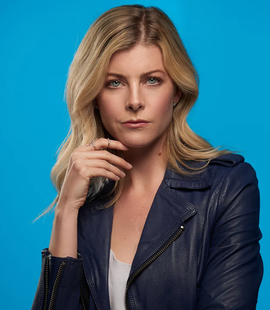 Actor headshot of a blonde woman in a navy leather jacket with her hand at her chin against a bright blue background