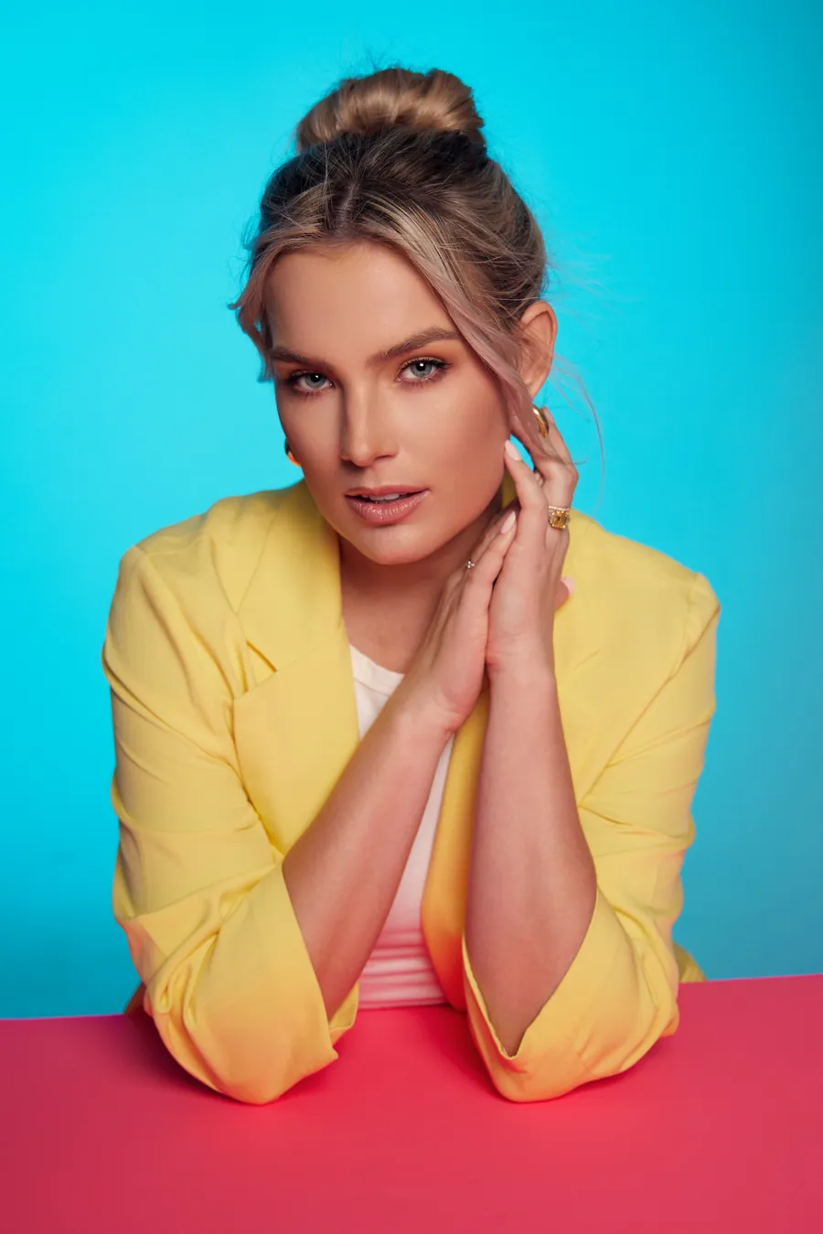 Actor headshot of a blonde woman in a yellow blazer with her hands together, against a cyan backdrop and a pink table