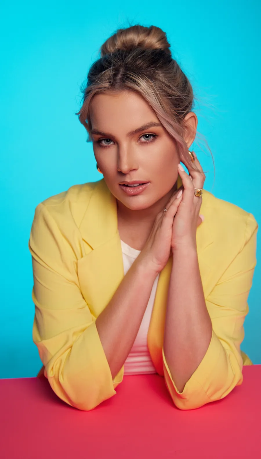 Actor headshot of a blonde woman in a yellow blazer with her hands together, against a cyan backdrop and a pink table