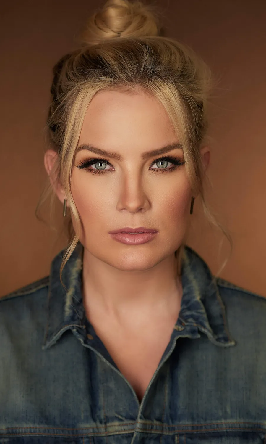 Actor headshot of a blonde woman with her hair in a bun, wearing a denim jacket against a warm brown background