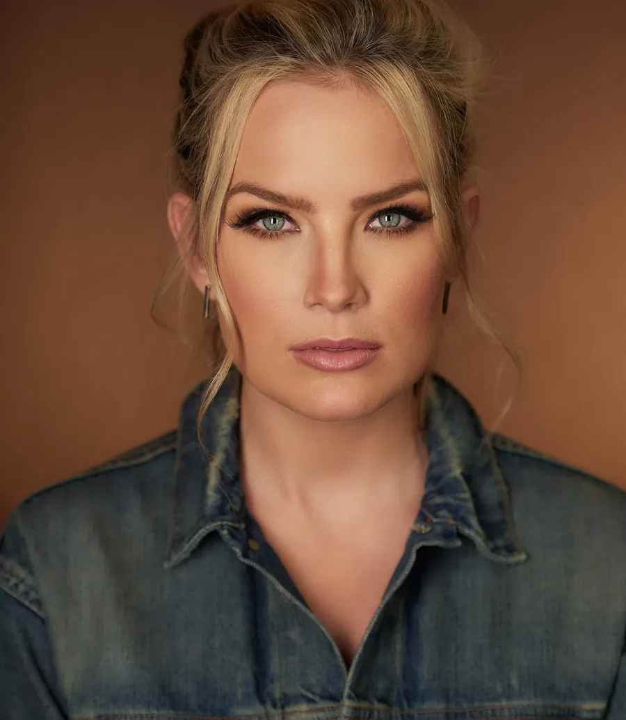Actor headshot of a blonde woman with her hair in a bun, wearing a denim jacket against a warm brown background