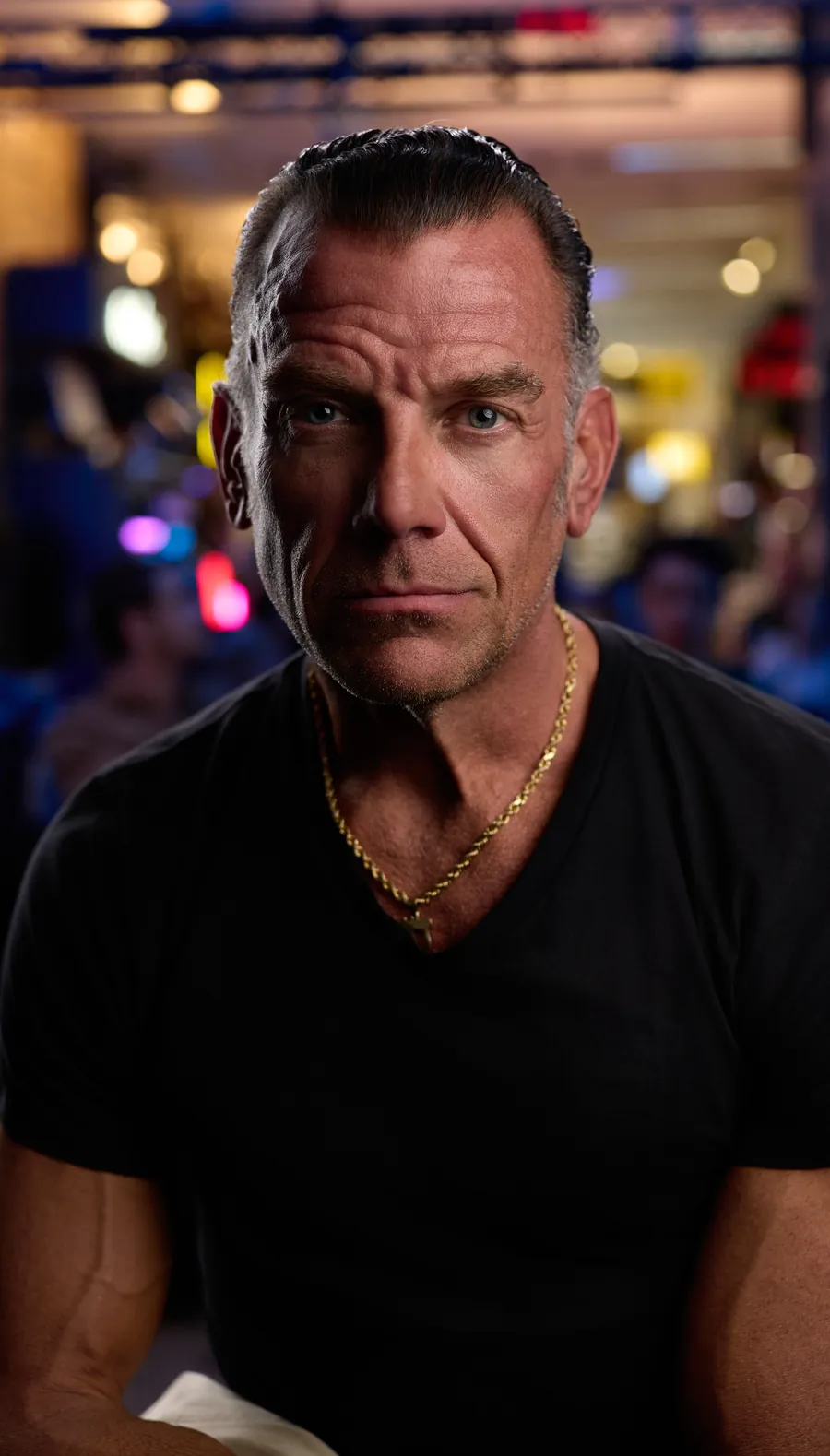 Actor headshot of a man with graying hair in a black t-shirt and gold chain, with colorful lights blurred behind him