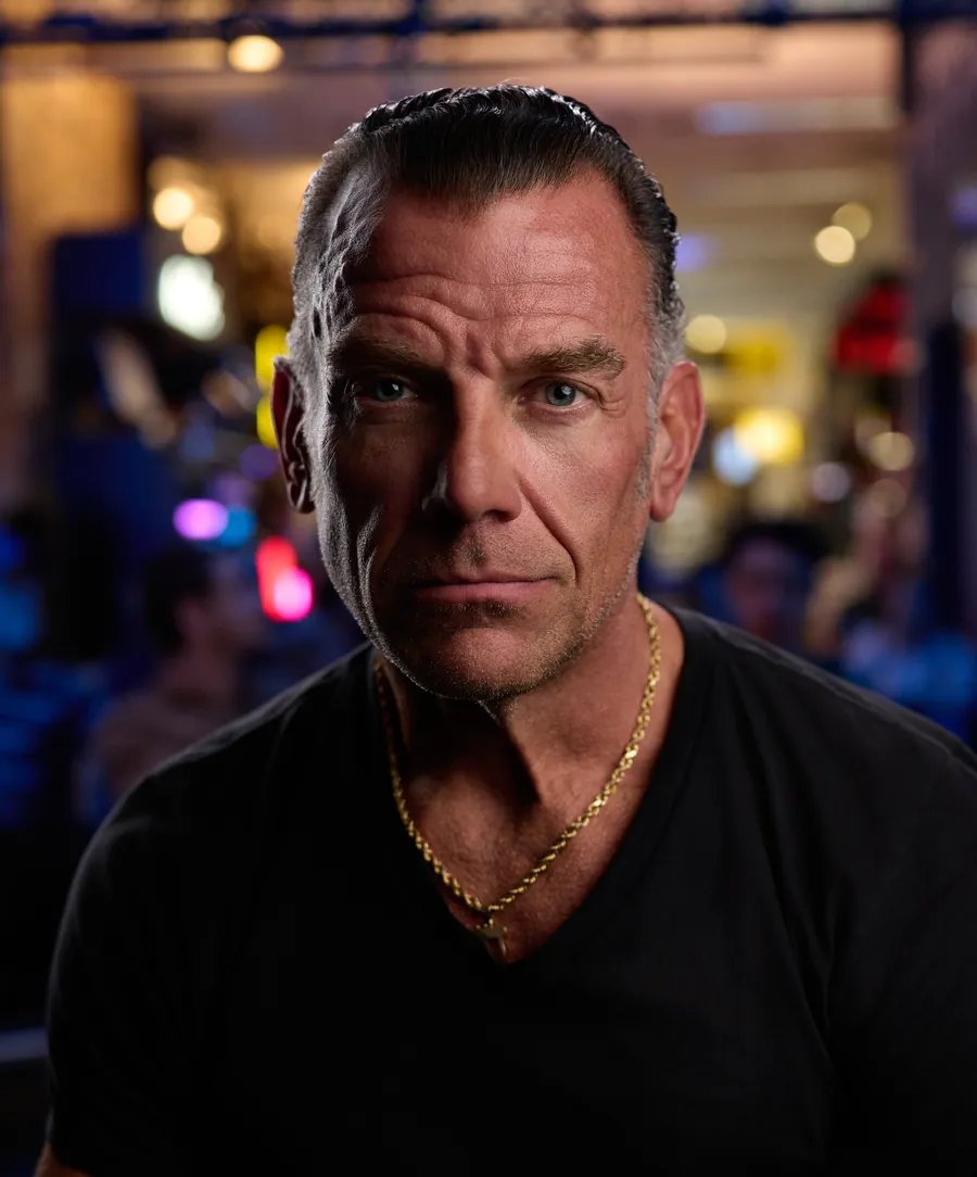 Actor headshot of a man with graying hair in a black t-shirt and gold chain, with colorful lights blurred behind him