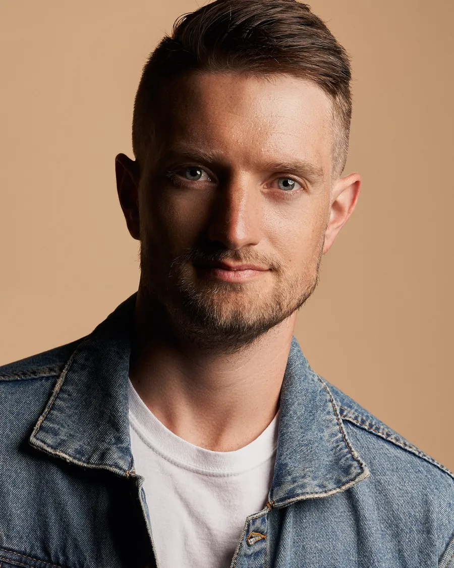 Actor headshot of a man with short swept hair and stubble in a denim jacket over a white t-shirt against a warm tan background