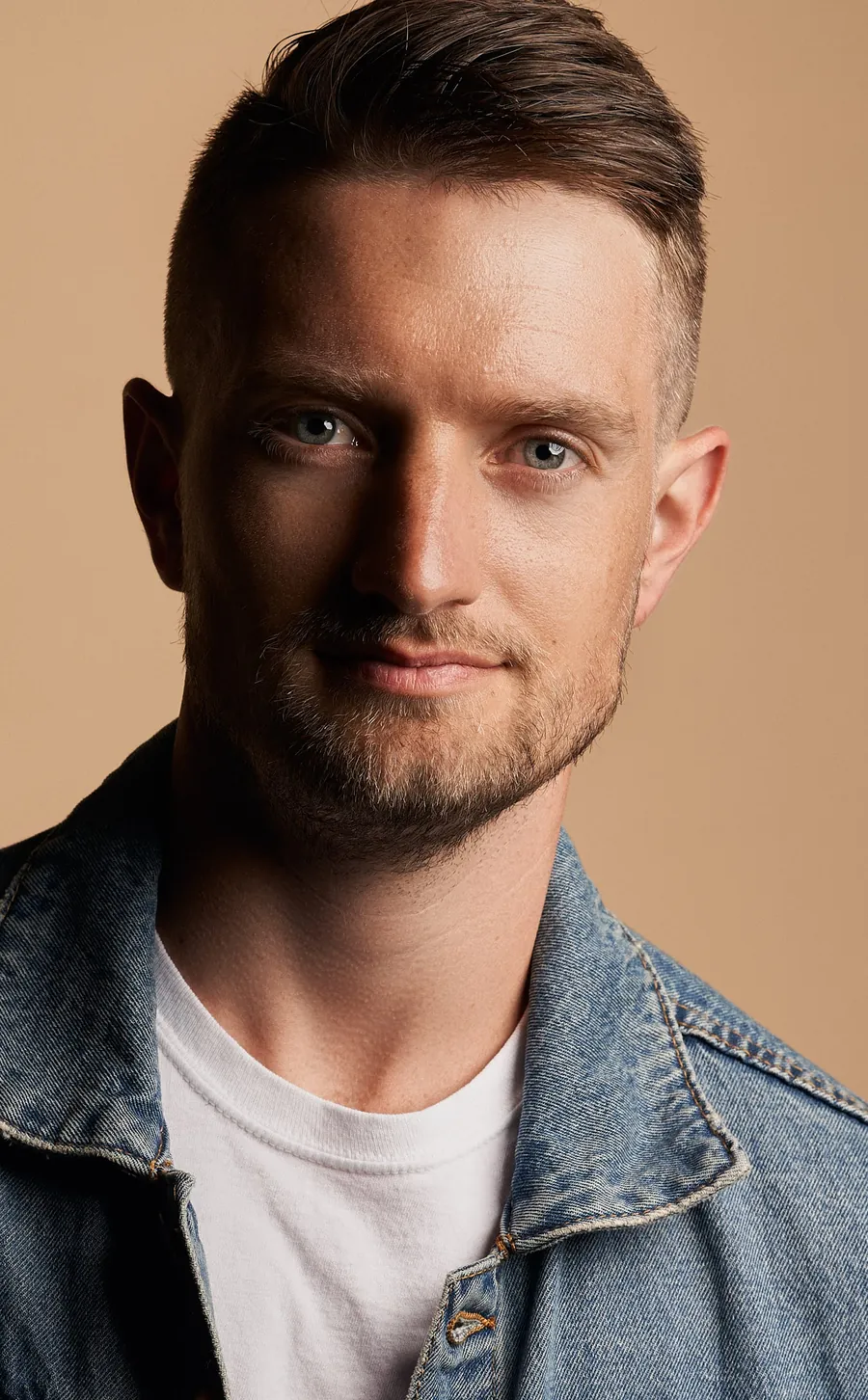 Actor headshot of a man with short swept hair and stubble in a denim jacket over a white t-shirt against a warm tan background