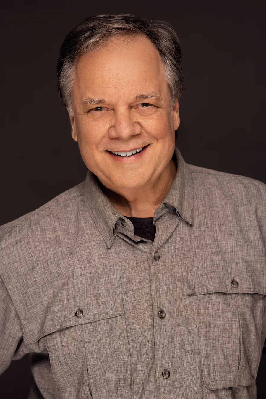 Actor headshot of a smiling older man with gray hair in a khaki button-up shirt against a dark brown background