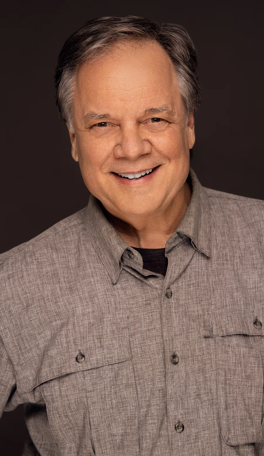 Actor headshot of a smiling older man with gray hair in a khaki button-up shirt against a dark brown background
