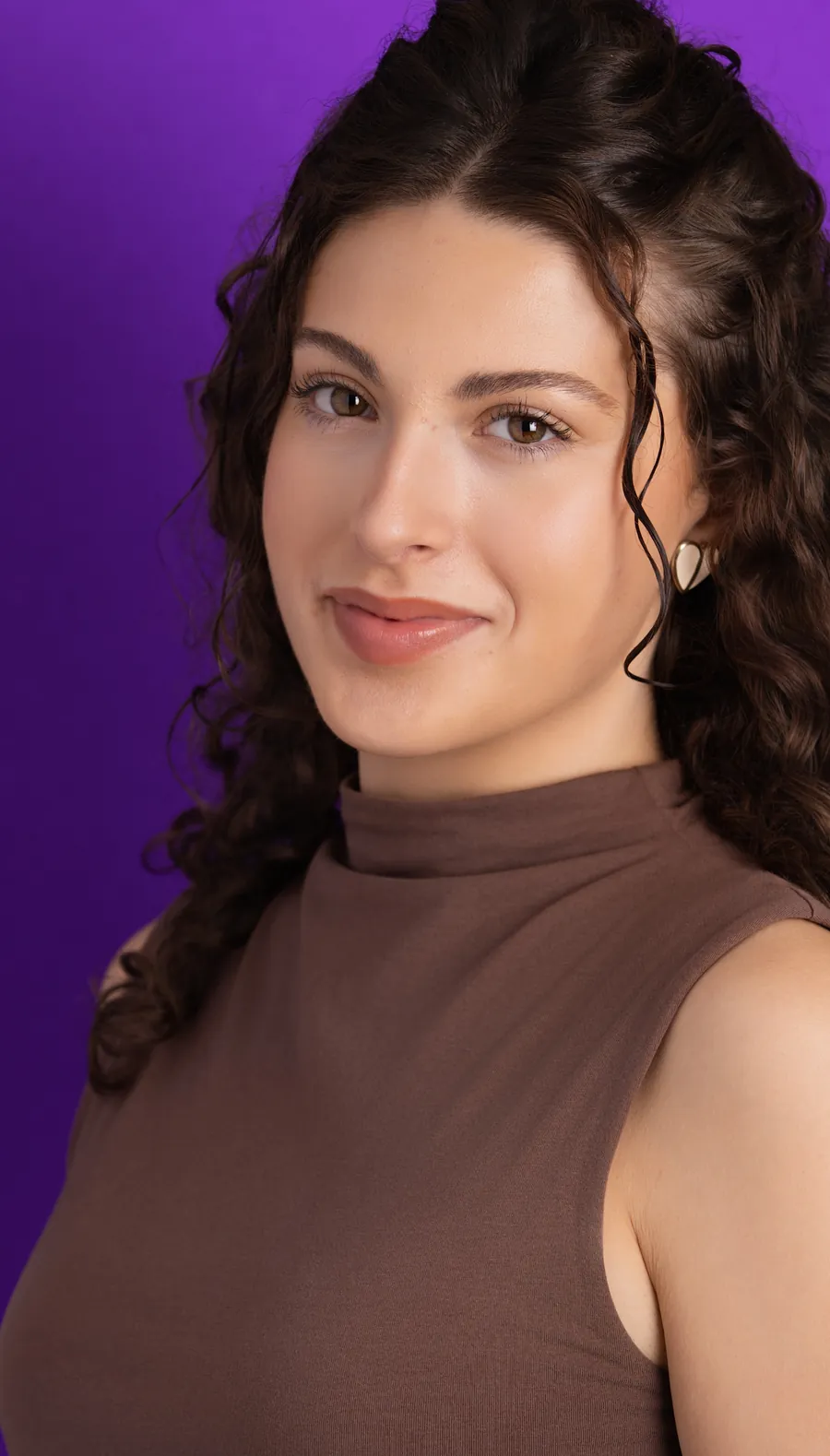 Actor headshot of a smiling woman with dark curly hair in a brown sleeveless top against a purple background