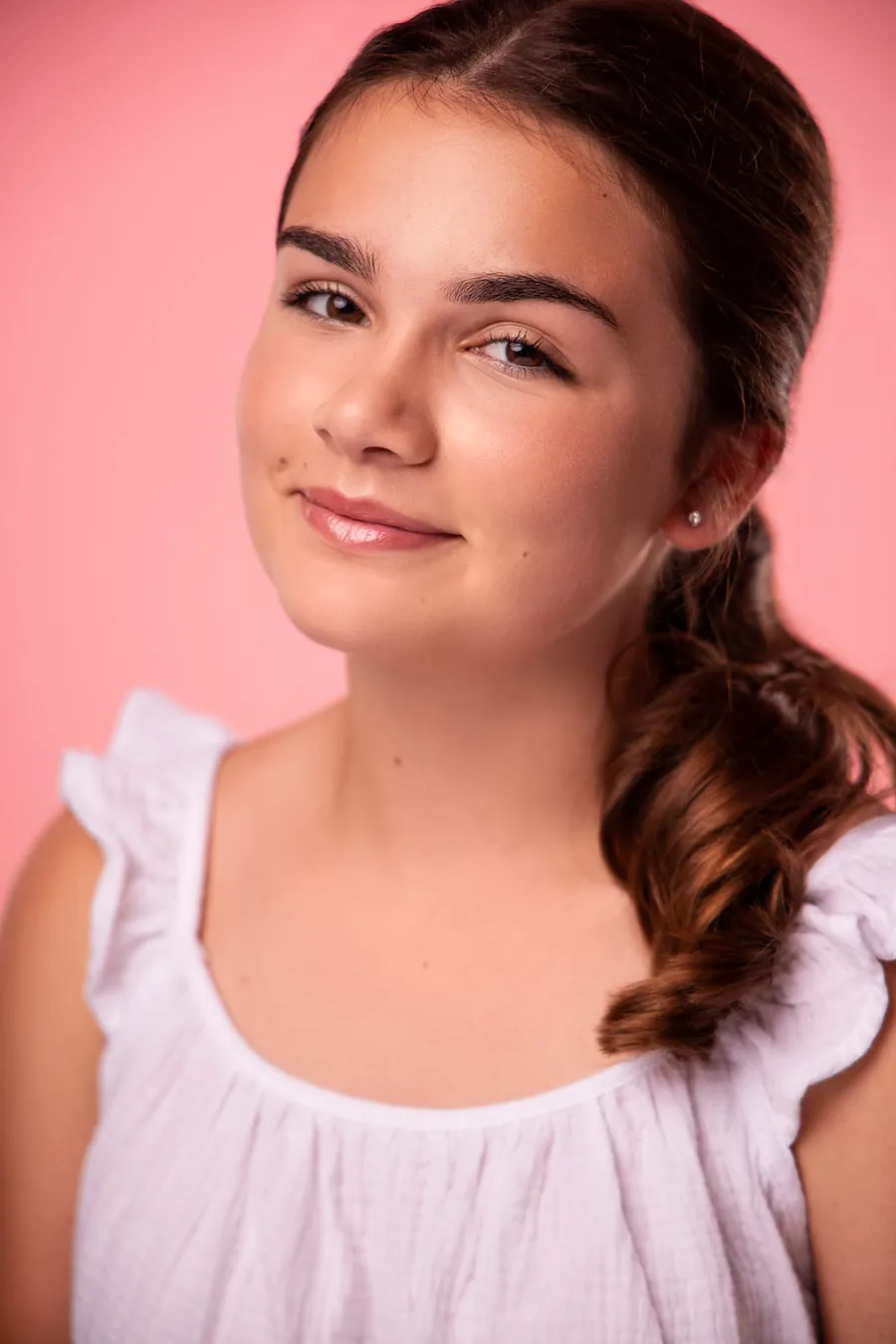 Actor headshot of a smiling young girl with a side ponytail in a white ruffled top against a pink background