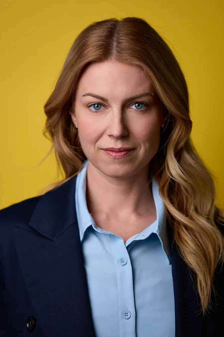 Actor headshot of a woman with long strawberry-blonde hair in a navy blazer and light blue shirt against a yellow background