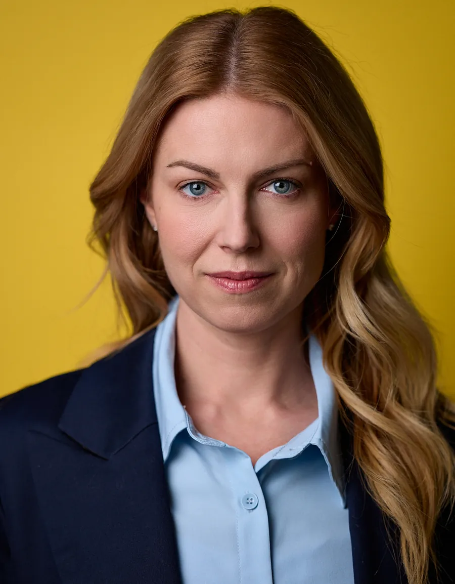 Actor headshot of a woman with long strawberry-blonde hair in a navy blazer and light blue shirt against a yellow background
