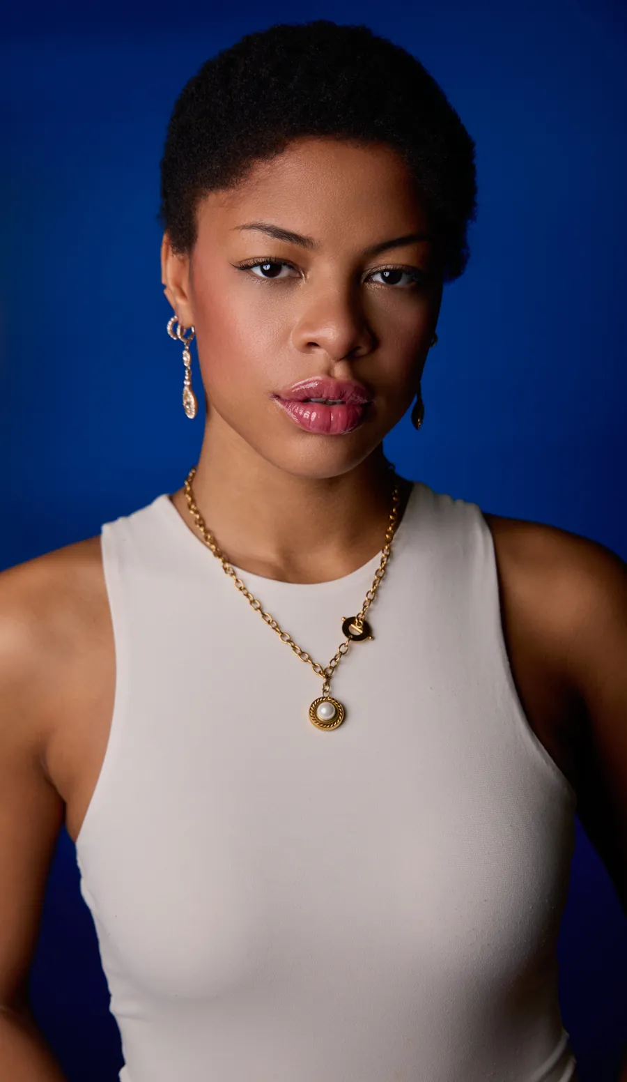 Actor headshot of a woman with short natural hair in a white tank top and gold necklace against a deep blue background