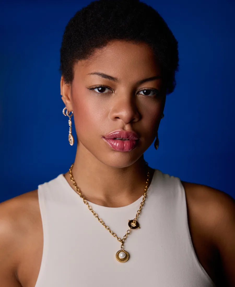 Actor headshot of a woman with short natural hair in a white tank top and gold necklace against a deep blue background