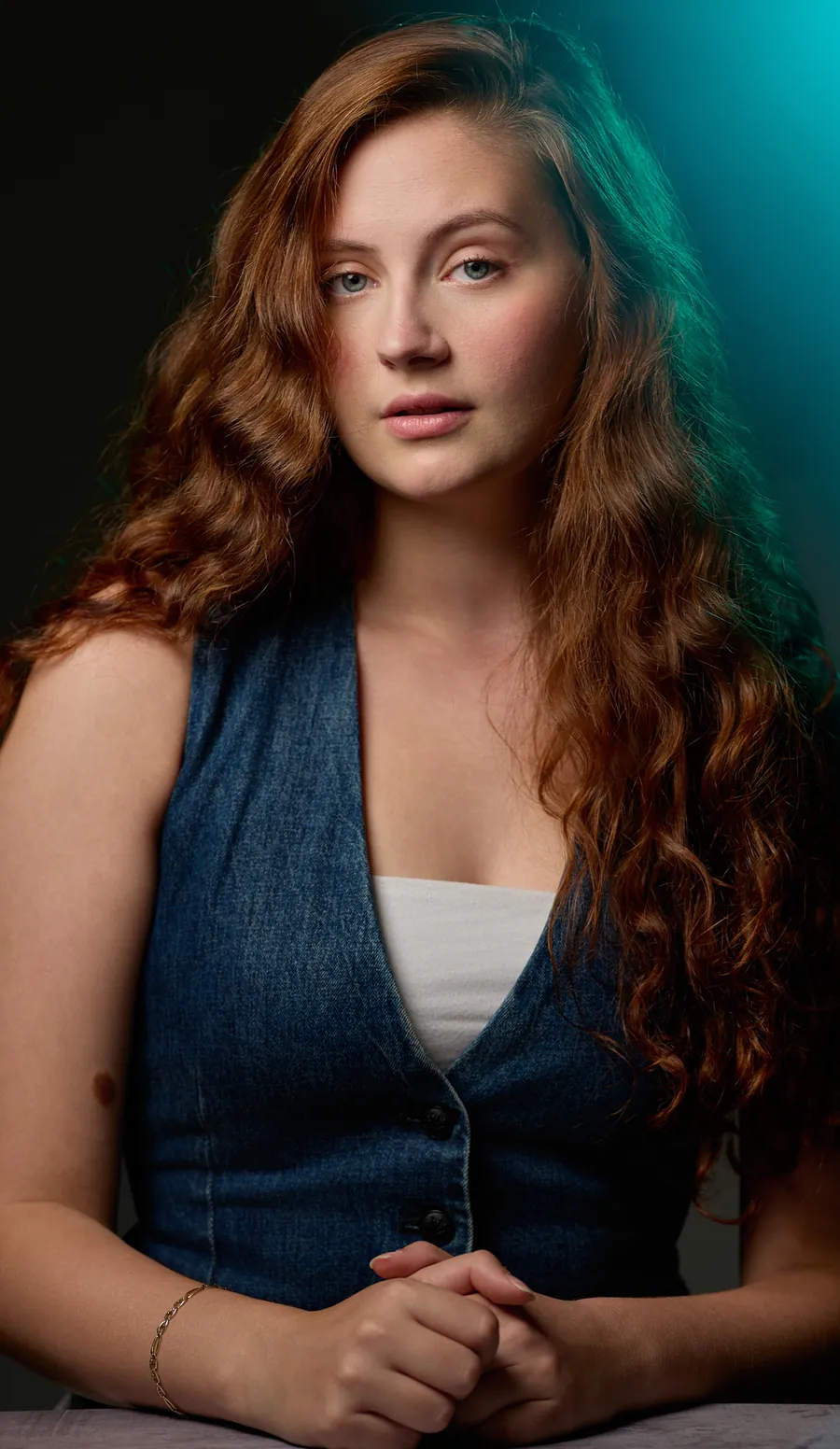 Actor headshot of a young woman with long auburn curls in a denim vest, lit with teal light against a dark background