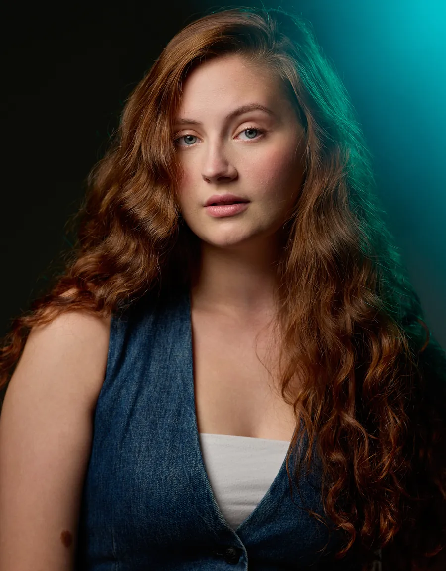 Actor headshot of a young woman with long auburn curls in a denim vest, lit with teal light against a dark background