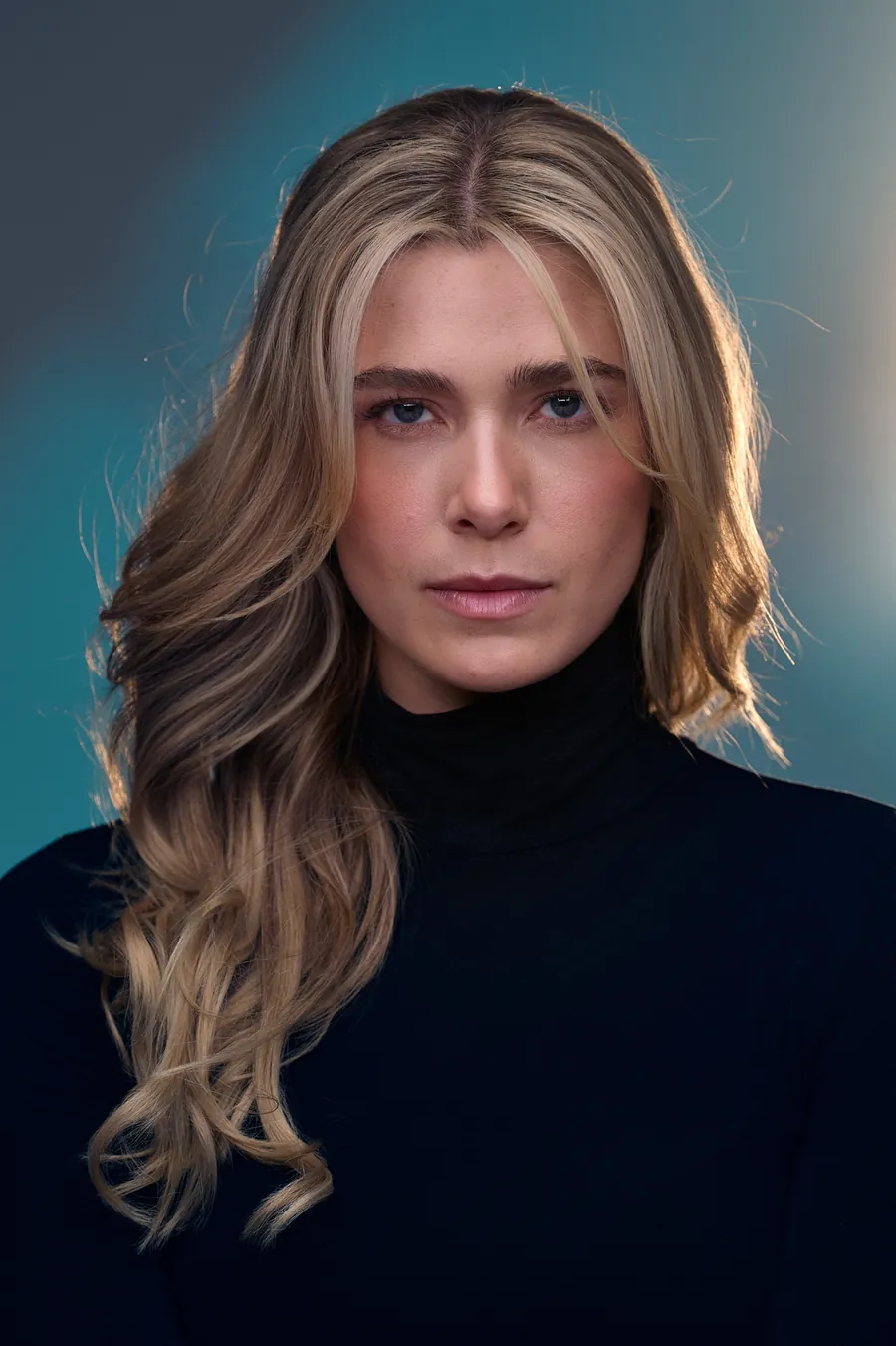 Actor headshot of a young woman with long blonde hair in a black turtleneck against a teal-gray backdrop