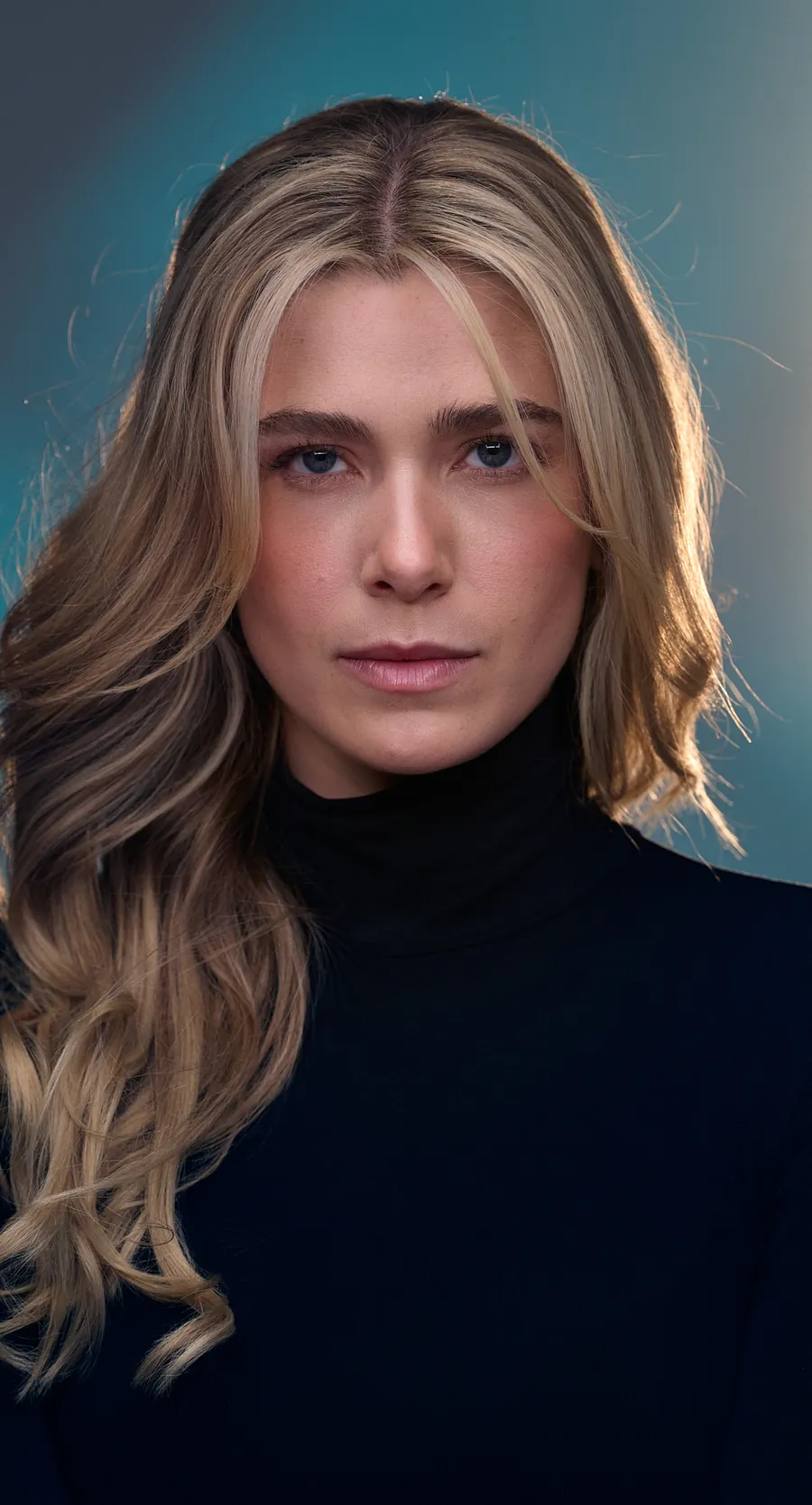 Actor headshot of a young woman with long blonde hair in a black turtleneck against a teal-gray backdrop