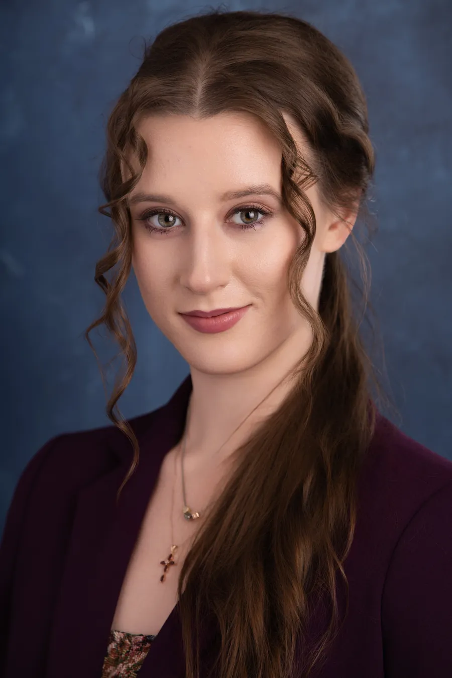 Actor headshot of a young woman with long braided brown hair and a burgundy top against a blue textured backdrop