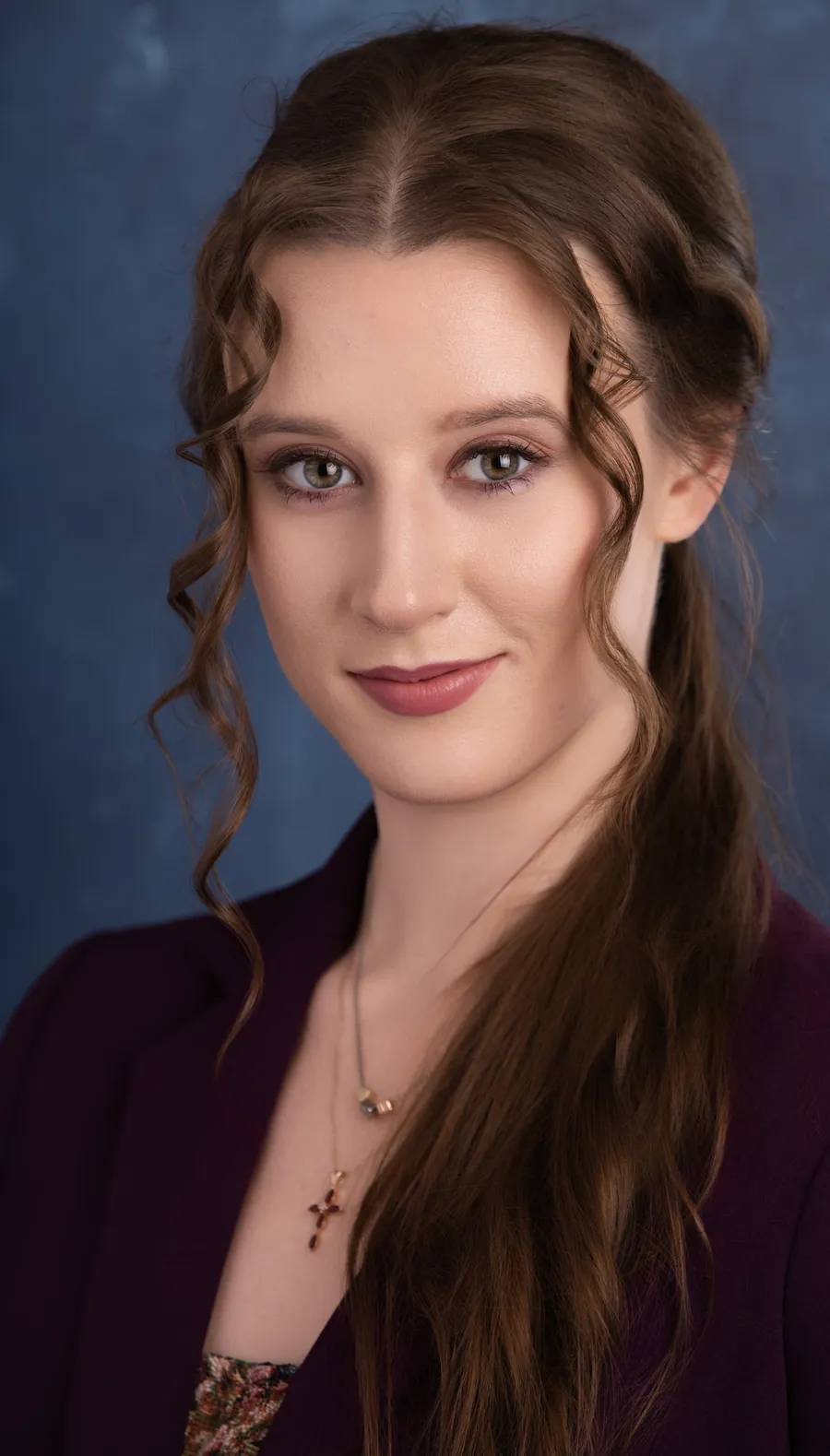 Actor headshot of a young woman with long braided brown hair and a burgundy top against a blue textured backdrop