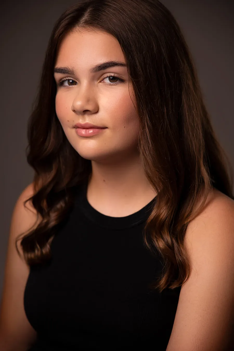 Actor headshot of a young woman with long dark wavy hair in a black tank top against a warm brown background