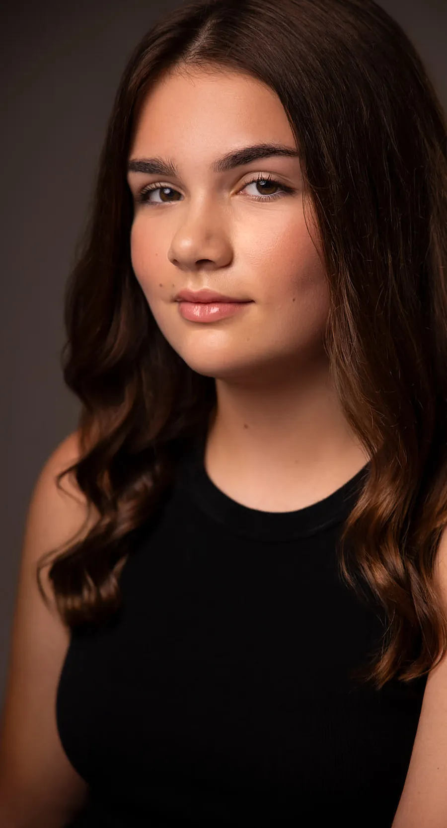 Actor headshot of a young woman with long dark wavy hair in a black tank top against a warm brown background