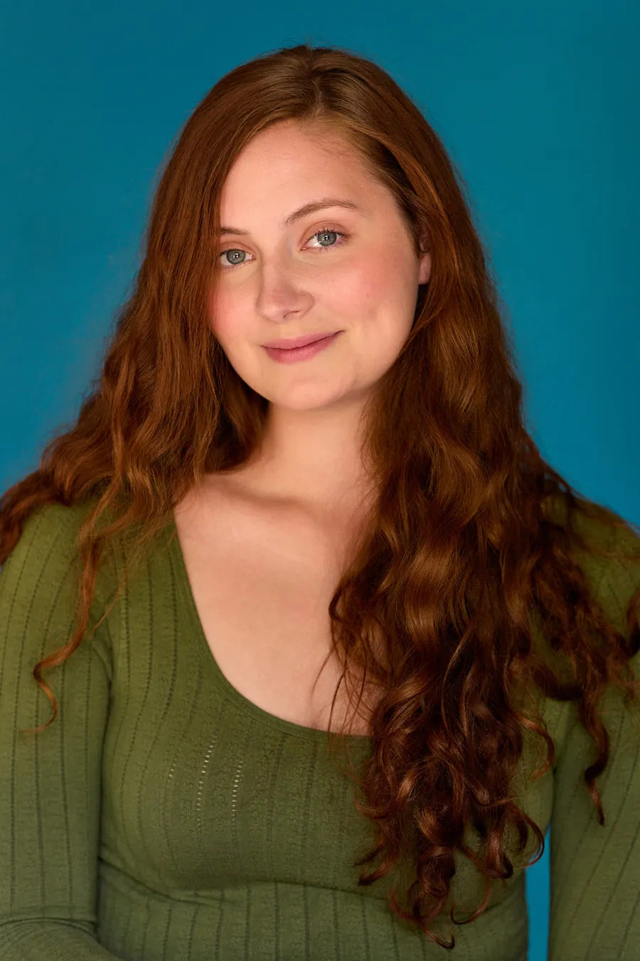 Actor headshot of a young woman with long red wavy hair in a green knit top against a teal backdrop
