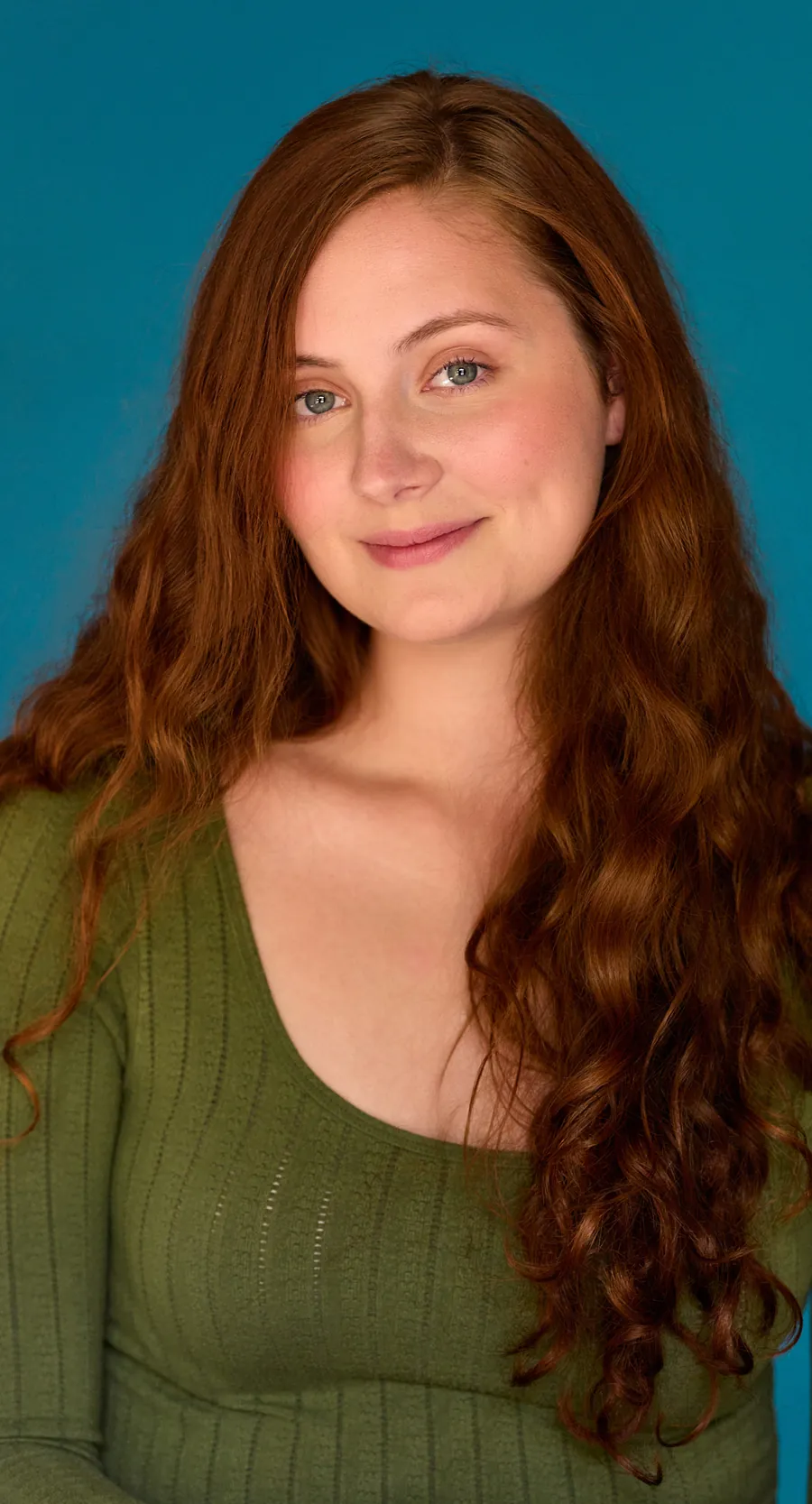 Actor headshot of a young woman with long red wavy hair in a green knit top against a teal backdrop