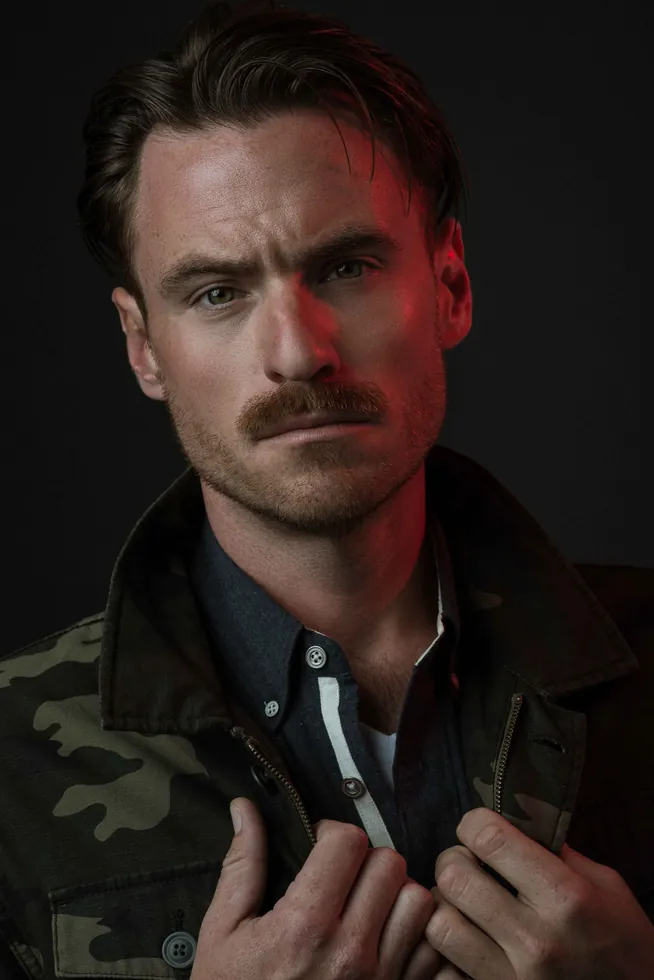 Character acting headshot of a man with a mustache in a camouflage jacket, lit with red light against a dark background