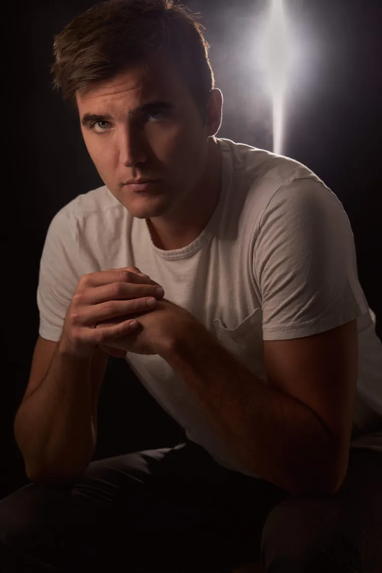 Character acting headshot of a man in a white t-shirt with clasped hands, lit from behind against a dark background