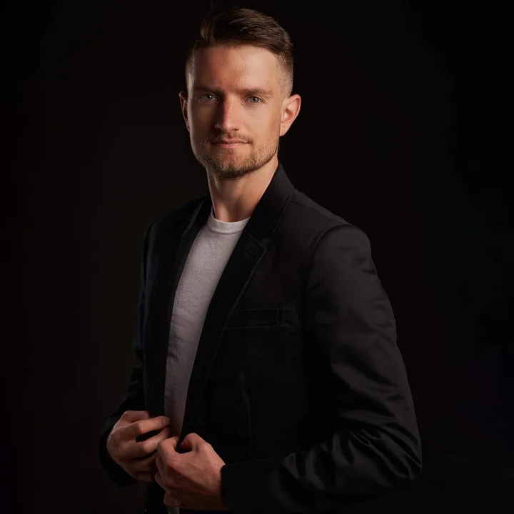 Character acting headshot of a man in a dark suit jacket over a white t-shirt adjusting his jacket against a black background