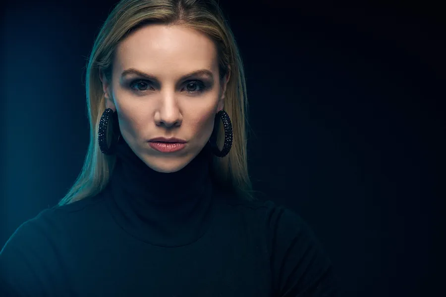 Cinematic actor headshot of a woman in a black turtleneck with large hoop earrings, lit in teal against a dark background