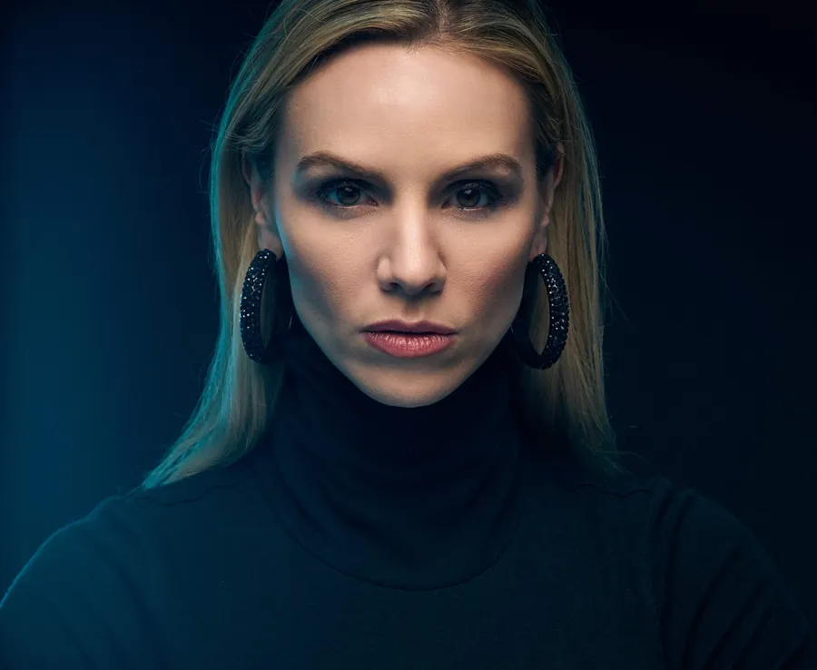 Cinematic actor headshot of a woman in a black turtleneck with large hoop earrings, lit in teal against a dark background