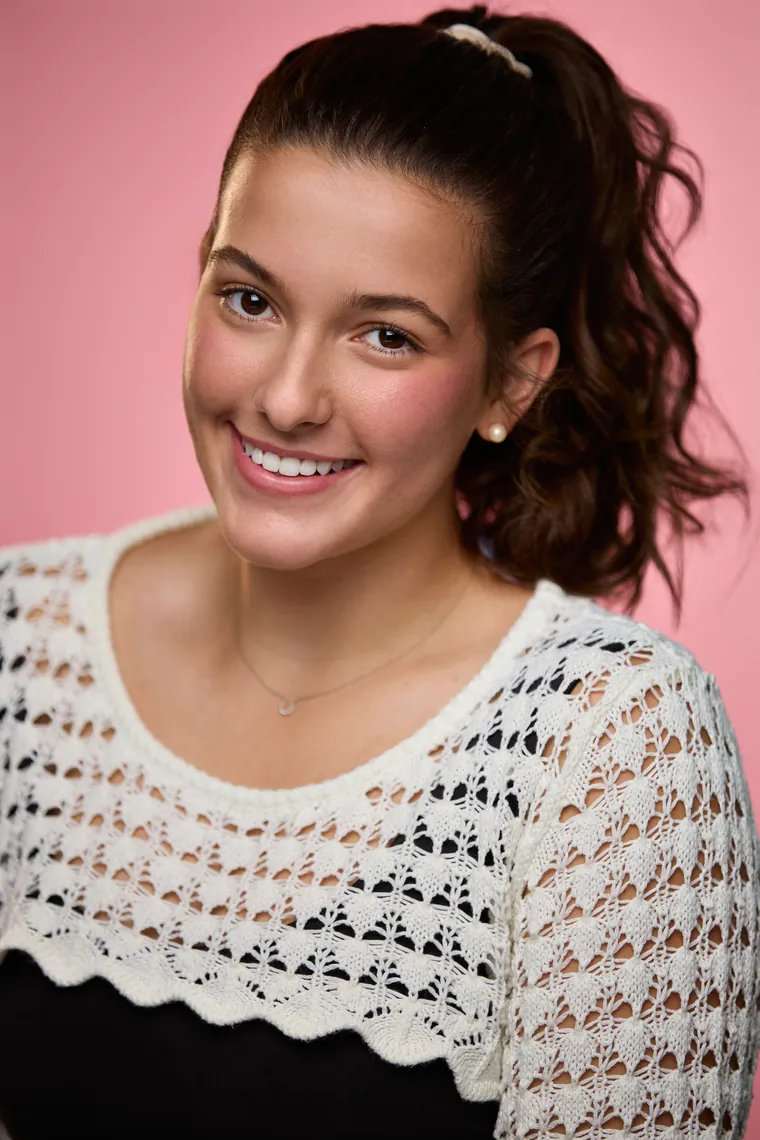 Commercial acting headshot of a smiling young woman with a high dark ponytail in a white knit top against a pink background