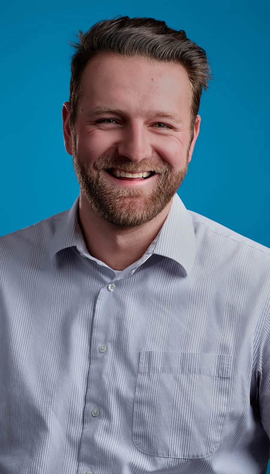 Commercial actor headshot of a smiling bearded man in a light blue button-up shirt against a bright blue background