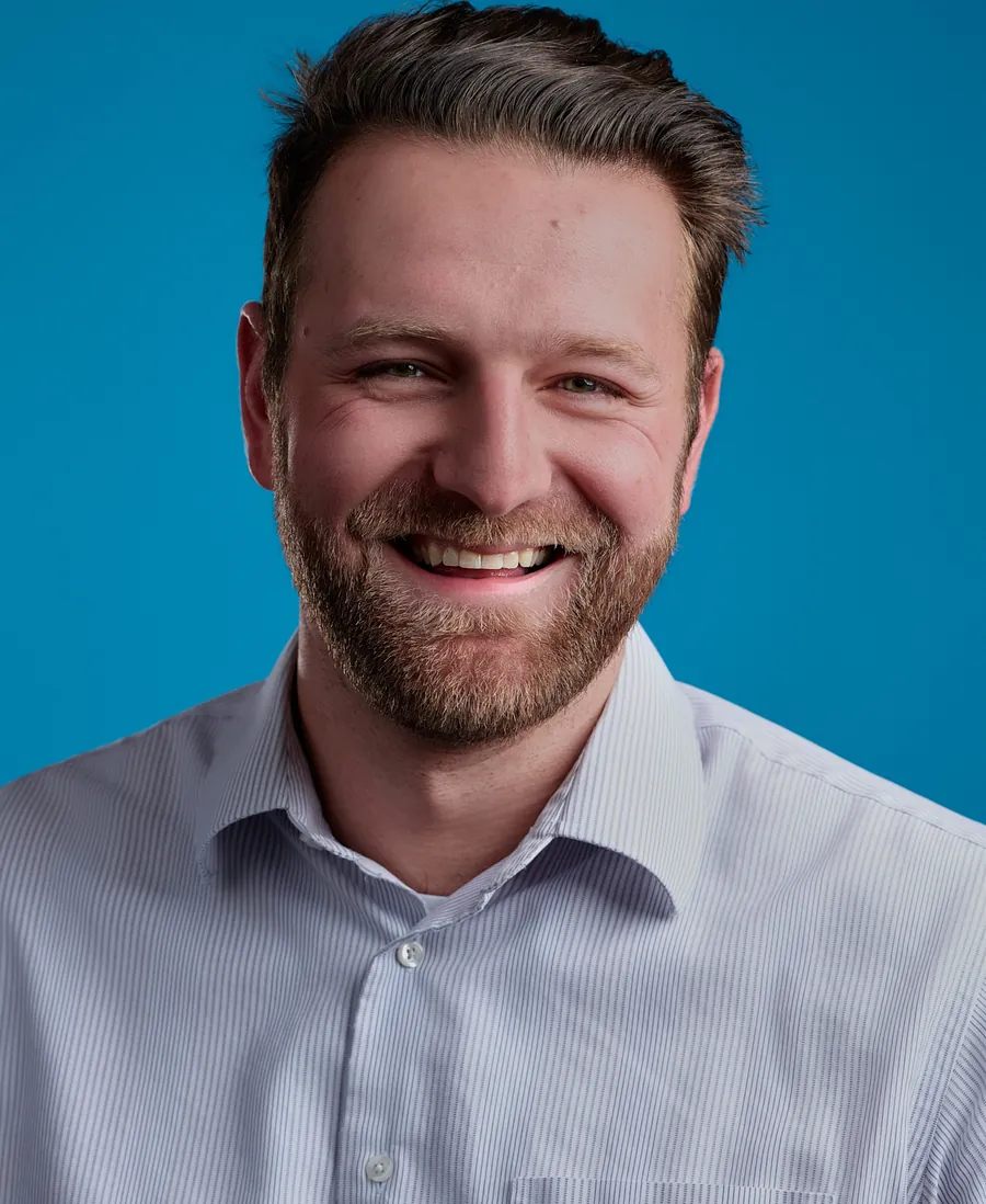 Commercial actor headshot of a smiling bearded man in a light blue button-up shirt against a bright blue background
