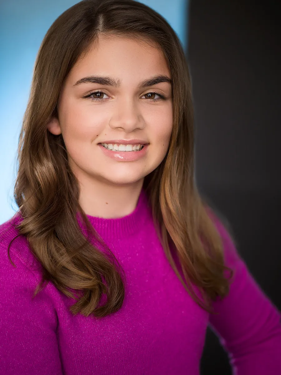 Commercial actor headshot of a smiling girl with long brown hair in a magenta sweater against a blue and dark backdrop
