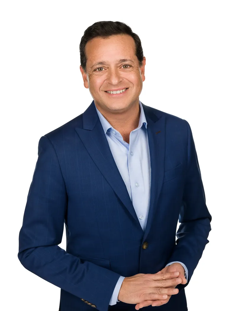 Smiling man in a blue suit and light blue shirt, hands clasped, against a plain white background