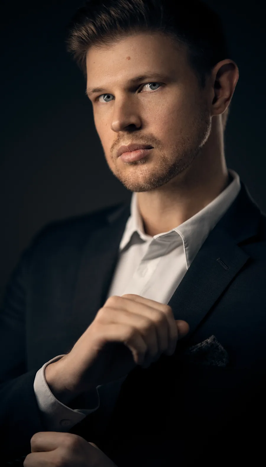 Dramatic actor headshot of a man in a dark suit adjusting his jacket, lit against a dark blue-black background