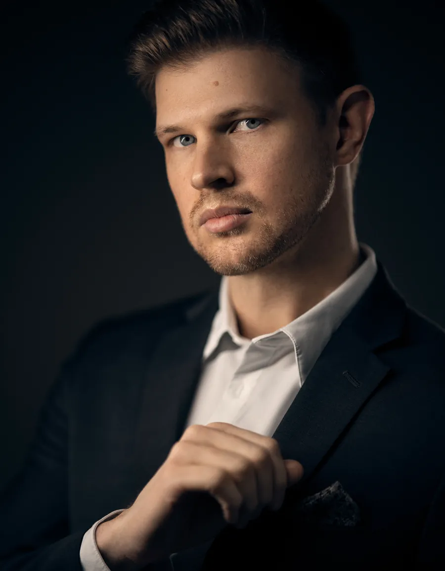 Dramatic actor headshot of a man in a dark suit adjusting his jacket, lit against a dark blue-black background