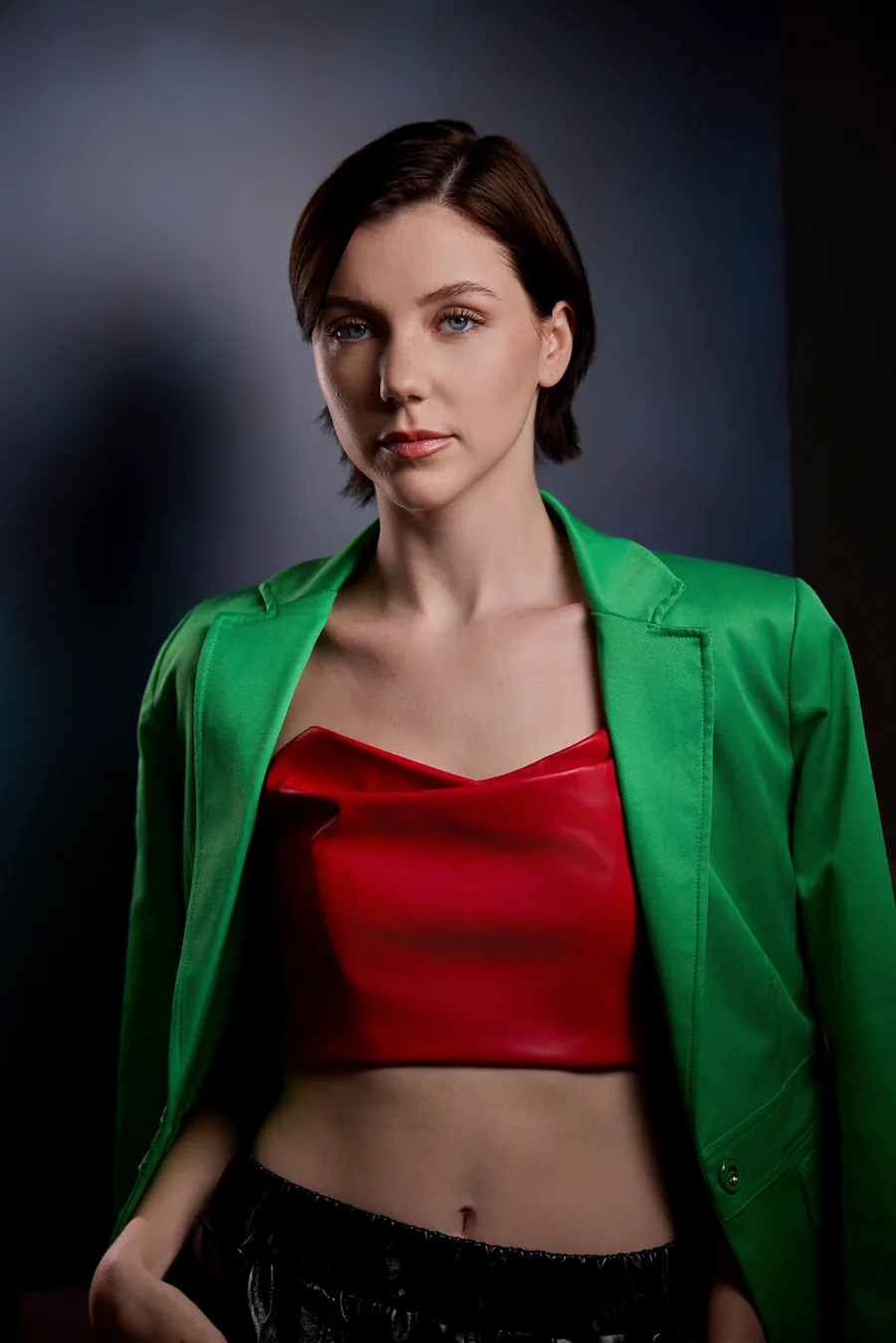 Editorial actor headshot of a woman with a short dark bob in a green blazer and red top against a gray studio backdrop