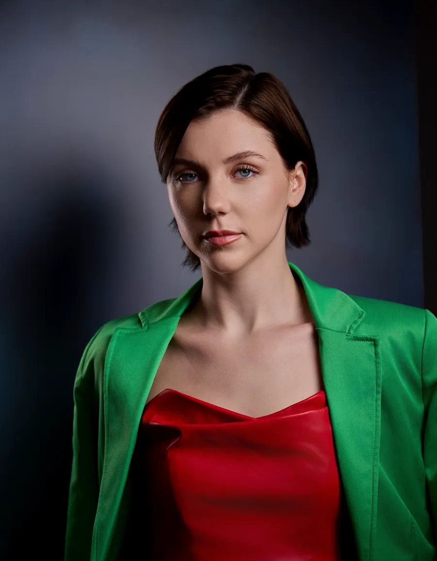 Editorial actor headshot of a woman with a short dark bob in a green blazer and red top against a gray studio backdrop