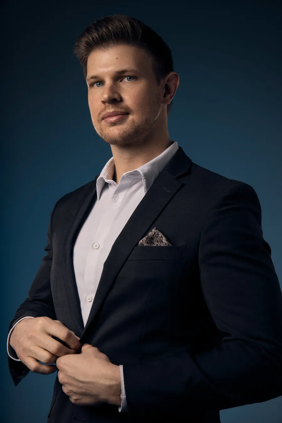 Editorial headshot of a man in a navy suit adjusting his jacket against a dark teal background