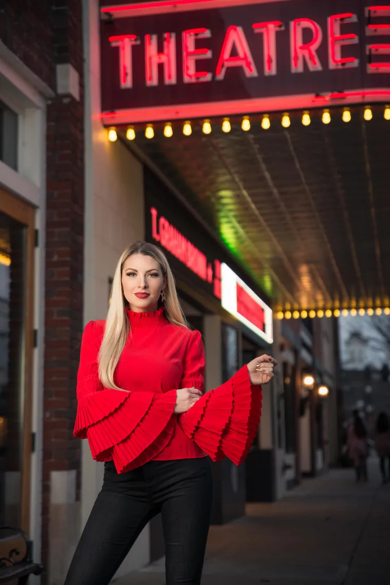 Woman in a red blouse with pleated sleeves posing confidently under a theater sign, lifestyle headshot
