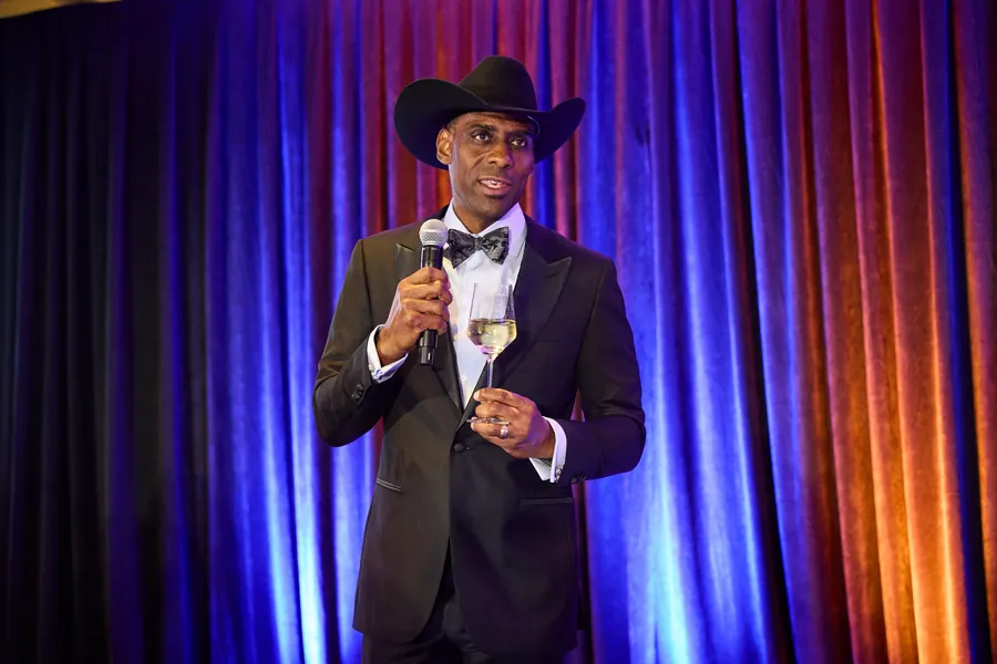 Man in a cowboy hat and tuxedo speaking into a microphone in front of a curtain