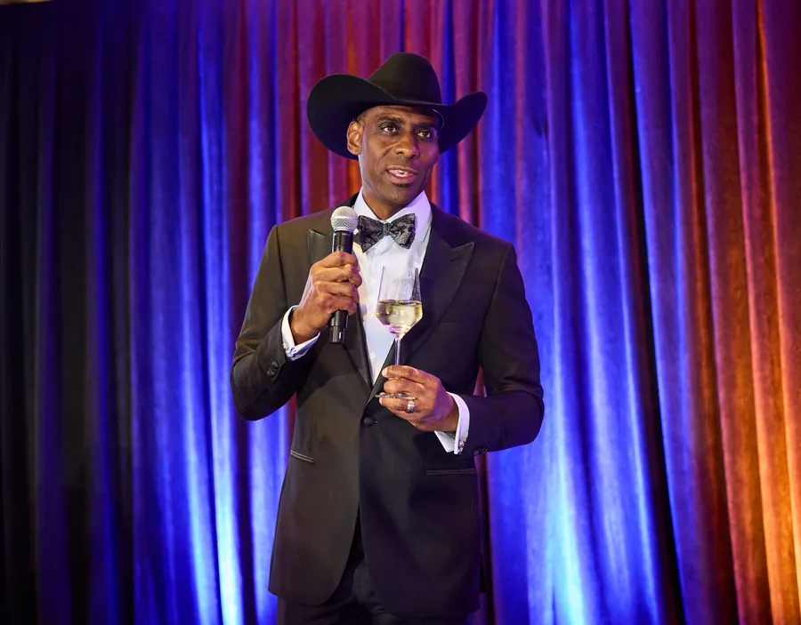 Man in a cowboy hat and tuxedo speaking into a microphone in front of a curtain