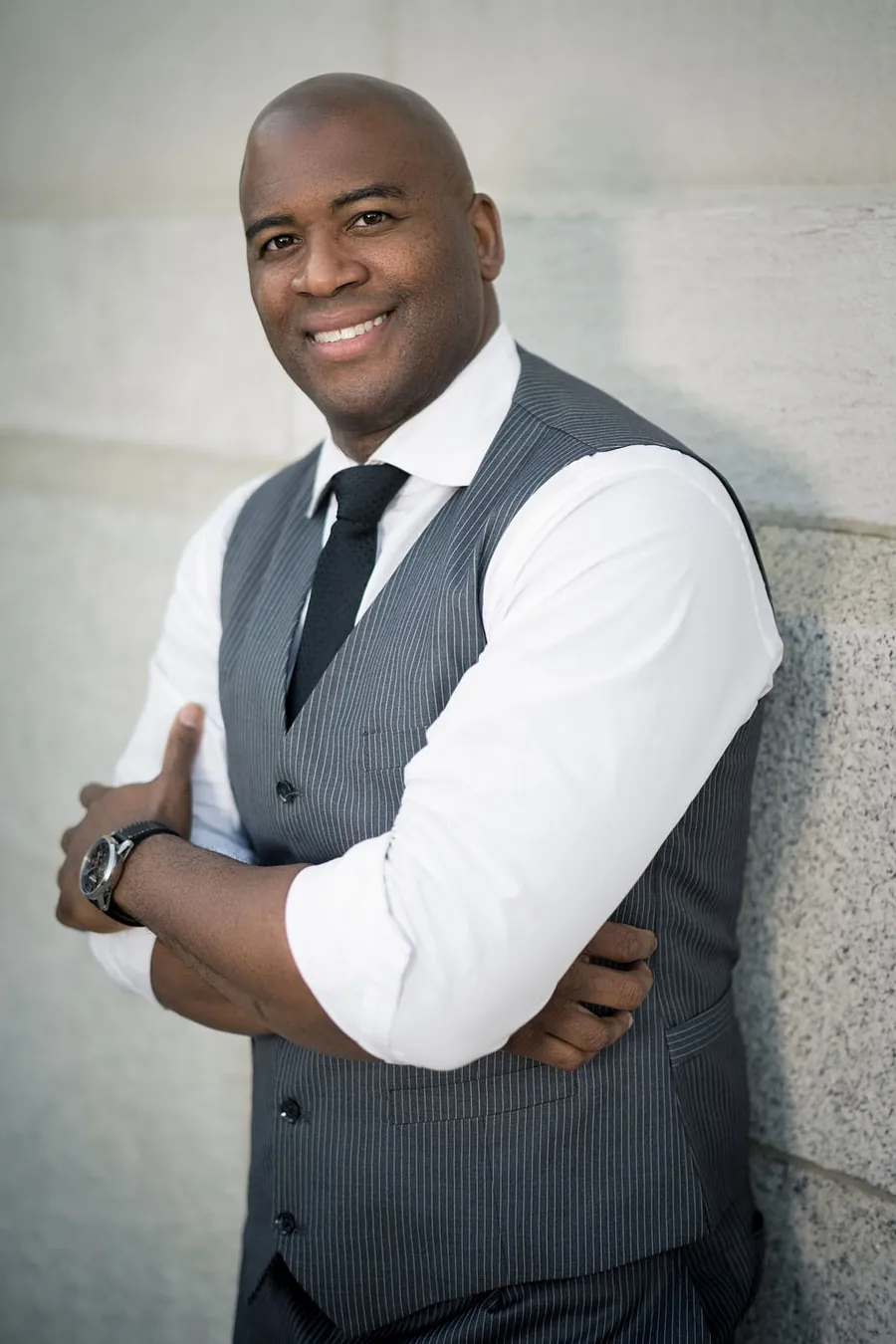 Man in a gray vest, white shirt, and tie with his arms crossed, leaning on a wall, business portrait