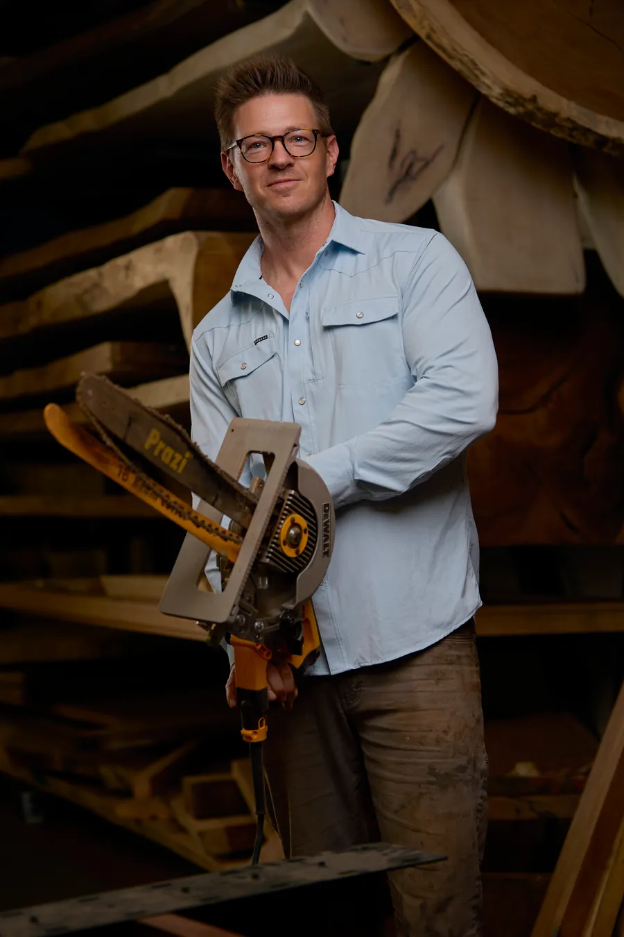 Man in a light blue shirt holding a chainsaw among wood slabs, branding portrait