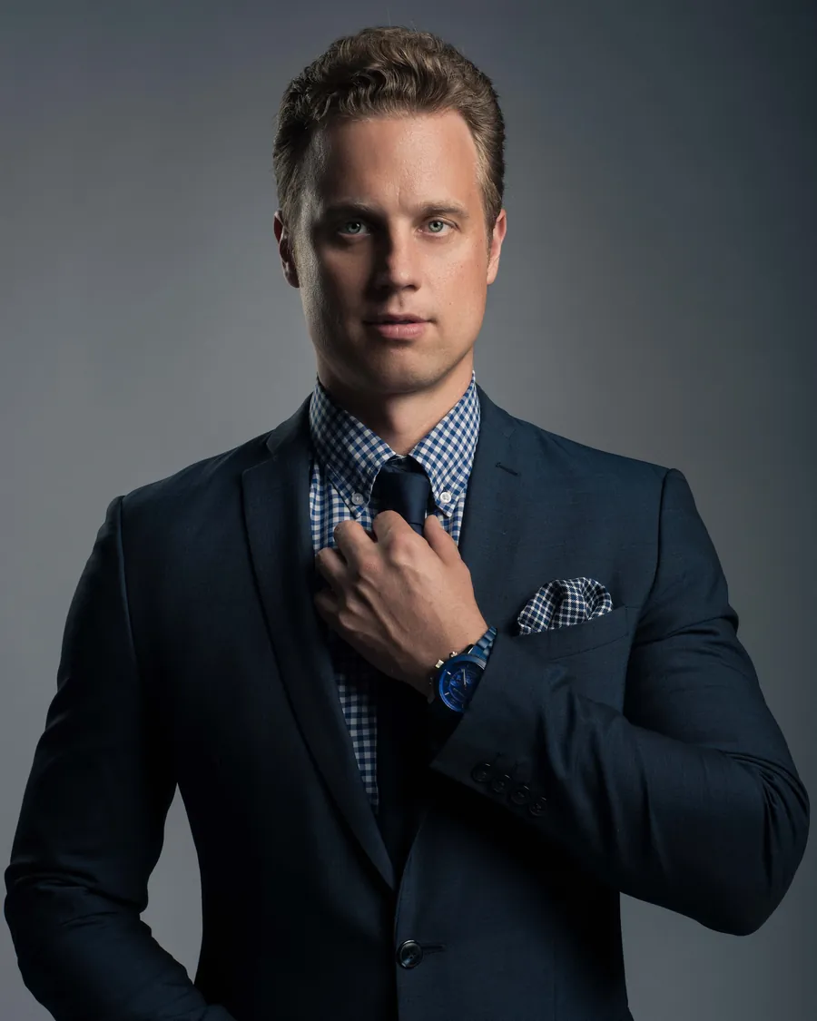 Man in a navy suit adjusting his tie against a moody gray studio backdrop, corporate headshot