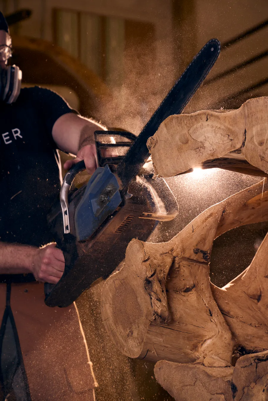 Man in a respirator cutting a large wood slab with a chainsaw, sawdust flying in the light