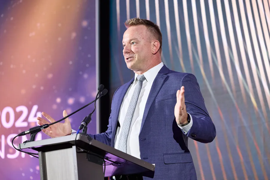 Man speaking at a podium in front of a patterned backdrop at a conference